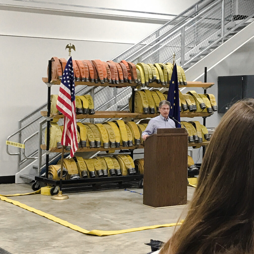 Man standing at a podium make a speech in a fire station with firehoses behind him
