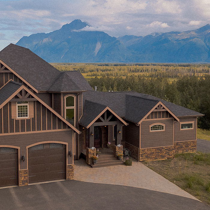 A large home with dark siding siting in front of a forest and mountains behind it